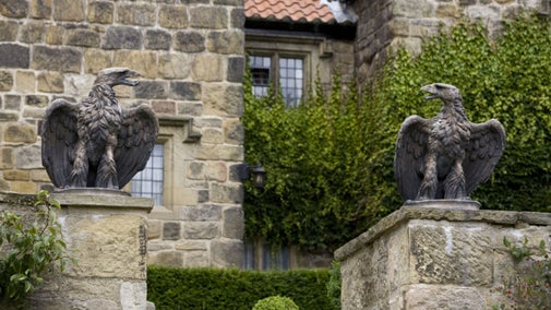 Two stone eagles sitting on top of stone pillars. A large stone building covered in ivy is in the background.